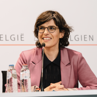 a woman with short dark hair , glasses and a pink jacket , sitting at the table and talking into the mic